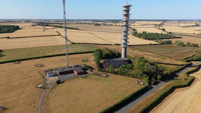 Aerial view of tall telecommunications transmitter mast surrounded by dry summer fields at sunrise in the UK