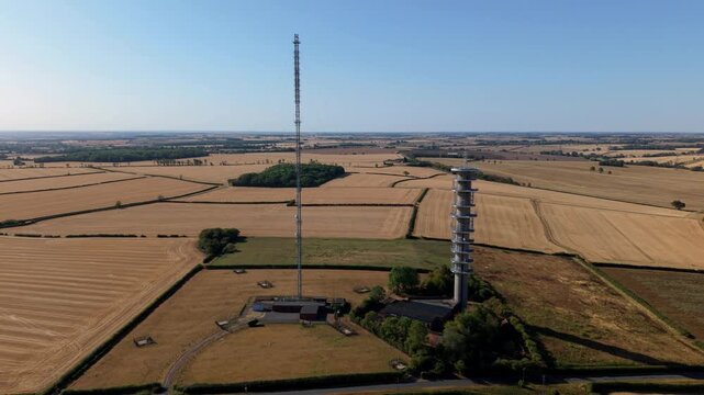 Aerial view of tall telecommunications transmitter mast surrounded by dry summer fields at sunrise in the UK