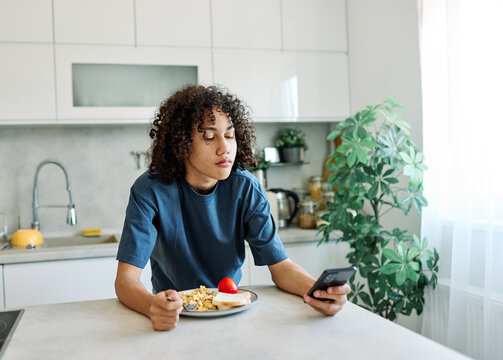 A teen sits at a kitchen counter eating breakfast and looking at a smartphone, balancing morning routine with digital distraction. The casual scene shows scrambled eggs and toast on a plate, curly hai