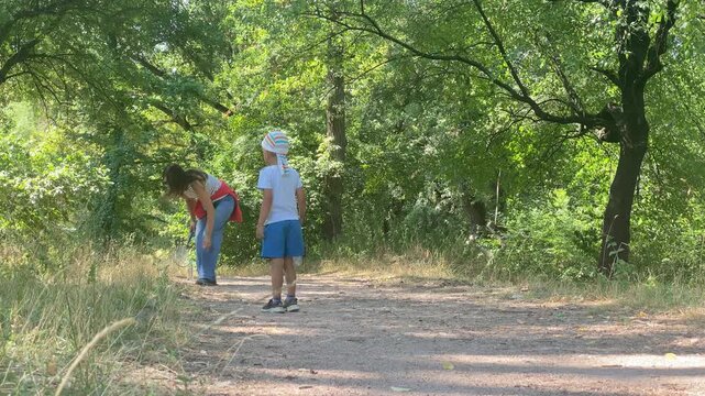 A mother bends down to pick up a shuttlecock while her young son stands watching. They play badminton on a shaded dirt path in a sunny forest. The peaceful scene evokes warm and joyful feelings.