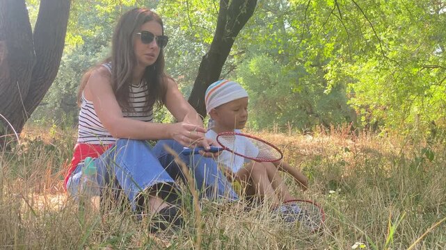 A mother rests thoughtfully while her young child plays with a shuttlecock. They sit together in dry grass under shady trees. The quiet moment brings a calm and peaceful feeling.