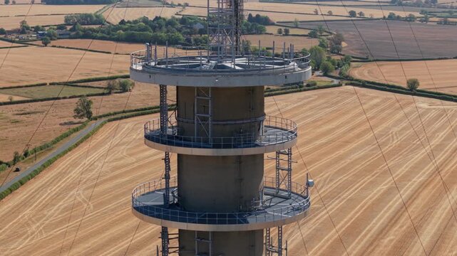 Close-up aerial view of the pinnacle and antenna array of a telecommunications transmitter mast over dry summer fields in the UK