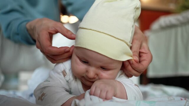 Careful mother's hands gently dressing a newborn baby with a soft yellow hat as the infant lies on the bed, preparing for the day and highlighting a moment of loving maternal care
