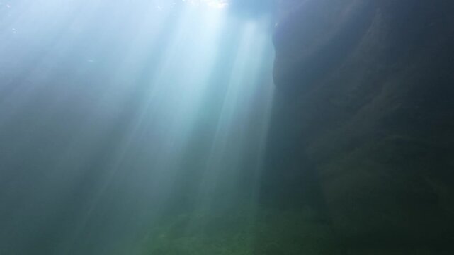 Broad sunbeams falling through the surface into a deep clear freshwater pool, lighting a dark rock wall and stony bottom in soft blue green, still and mysterious underwater scene in natural light