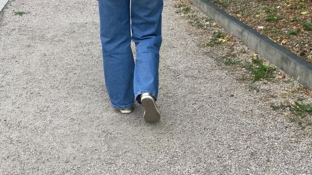 A woman wearing blue jeans walks calmly down a gravel path. Her gentle steps create a peaceful moment in the park. She enjoys a relaxed and carefree walk.