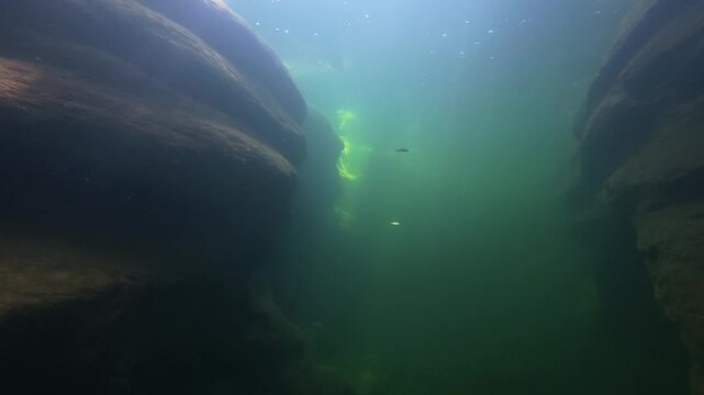 Underwater canyon of dark rock walls in a deep river pool, narrow sunbeams falling onto a bed of smooth pebbles and large boulders in cool blue green water, calm and mysterious freshwater scene