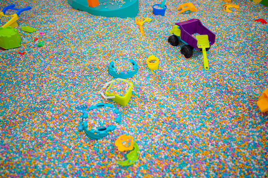 Various colorful plastic toys and shovels scattered in a sensory pit filled with multi colored foam beads at an indoor kids playground.