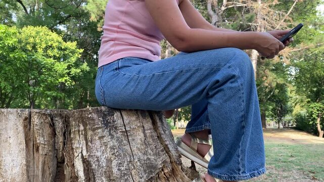 A woman sits comfortably on a large tree stump while holding her phone. She rests peacefully in a shady park surrounded by green trees. Her calm posture reflects quiet contentment.