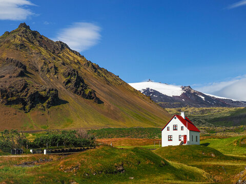 Atlantic Ocean shore, Faxafloi Bay, with green meadows, a white prefect house, and the white, snowy peak of Snaefellsjokull Volcano, against the blue sky. Arnarstapi, Snaefellsnes Peninsula, Iceland.
