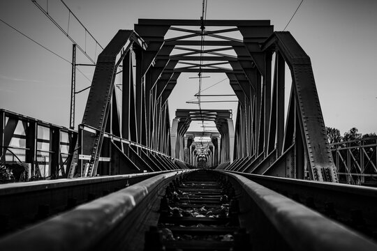 Black and white low angle view of railway tracks leading through a massive steel truss bridge structure in symmetrical perspective.