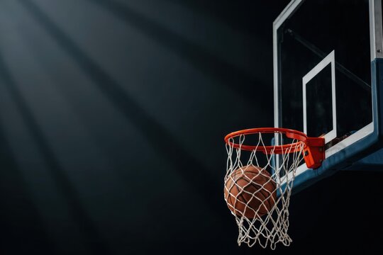 A basketball falling through the net on a hoop against a dark moody background with dramatic light beams. Perfect for sports and action concepts.