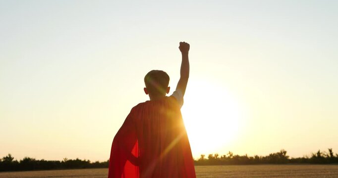 Young superhero boy raises fist at sunset in open field. Silhouette shows courage, hope, and imagination. Inspiring footage represents childhood dreams, strength, freedom, and empowerment.