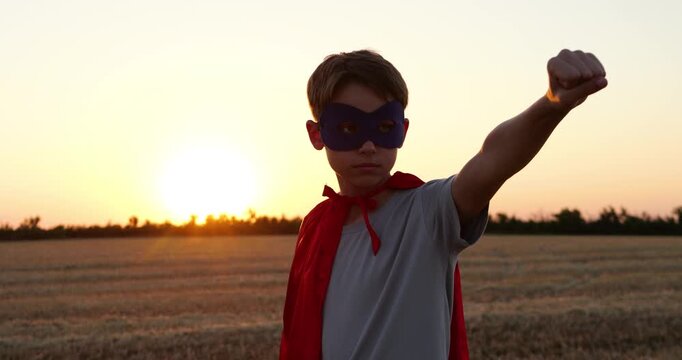Young superhero boy wears mask and red cape. Kid strikes heroic pose with raised fist in wheat field at sunset. Childhood imagination, courage, and adventure inspire fearless dreams.