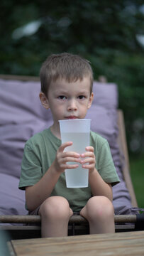Cute focused child quenches thirst with water outdoors on patio surrounded by greenery and lounge furniture. 