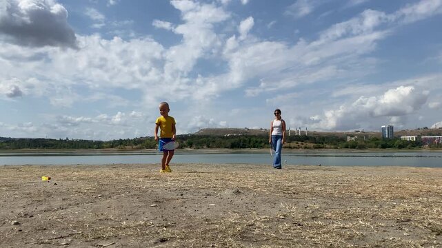 A young boy and a woman play a fun game near a scenic lake shore. They enjoy a cheerful day outdoors under a cloudy sky. The playful moment brings joy and family warmth.