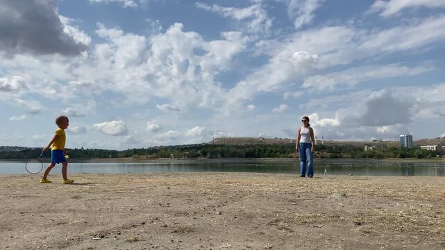 A young boy runs joyfully across the shore while holding a racket as his mother watches. They play near a peaceful lake under a cloudy sky. The playful moment brings happiness.