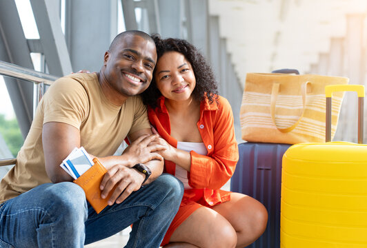 Smiling young Black couple sitting closely together beside colorful luggage in an airport terminal, holding passports and boarding passes while waiting for their holiday flight
