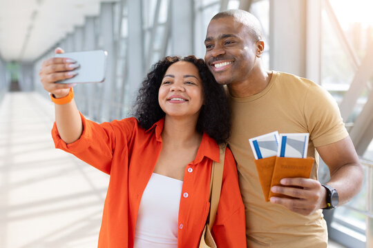 Young Black couple taking cheerful smartphone selfie at the airport while holding passports and boarding passes, capturing happy moment before their vacation flight