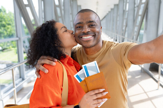 Young Black woman kissing her smiling partner while they take selfie at the airport, holding passports and boarding passes before departing together on a romantic trip