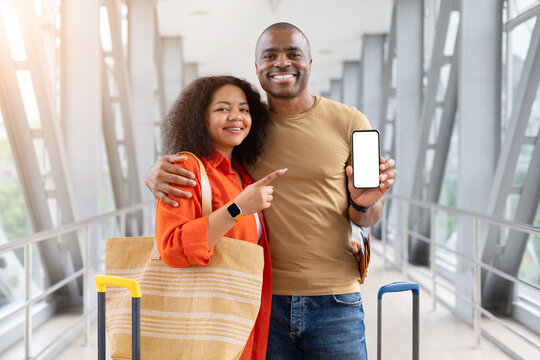 Smiling couple embracing at airport, man displaying blank smartphone screen while woman points at it, mockup template with luggage