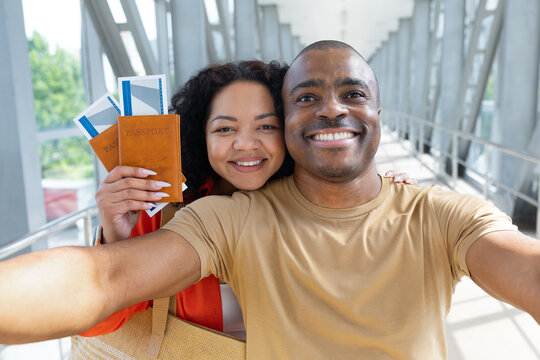 Young Black couple taking cheerful selfie in airport terminal while holding passports and boarding passes, capturing a happy travel moment before their vacation flight