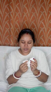 Indian young woman sick, suffering from fever and cold flu on sitting sofa, blowing on her nose with napkin or tissue, Pollen allergy, sneezing, flu fever concept.