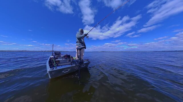 Fisherman is fishing on lake from boat casting spinning rod at daylight. Angler is standing in an equipped boat and catching fish while trolling. Man holds spinning rod while offshore during sunny day