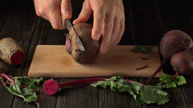 Hands slice a fresh beetroot on a wooden cutting board, revealing vivid red rings beside beet greens and whole roots on a dark rustic table