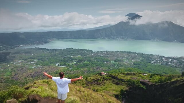 Man standing on a volcanic mountain peak with arms raised, celebrating success and freedom while overlooking Batur Lake and surrounding caldera landscape in Bali, Indonesia. Aerial view drone flight
