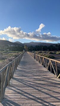 Vertical static view of wooden boardwalk with long shadows leading toward palm trees and mountains in Morro Jable, Fuerteventura