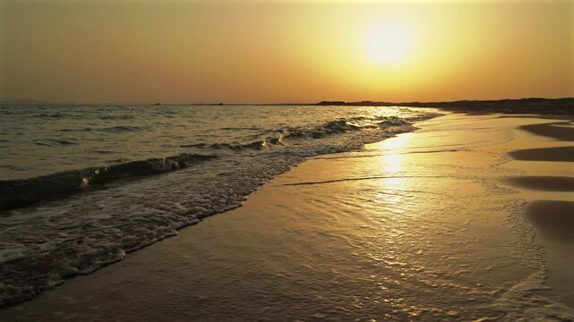 Wide-angle shot of golden sunset over calm ocean waves washing onto the sandy beach shore