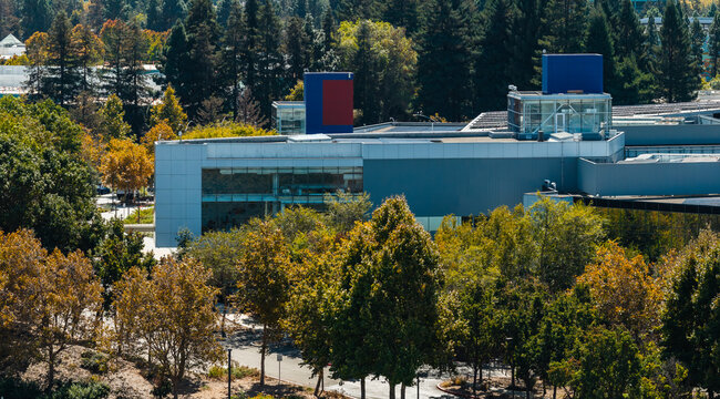Aerial perspective shows a modern corporate tech Google office building bordered by autumn trees, paved pathways, and glass architecture at a business park complex in Mountain View, California.