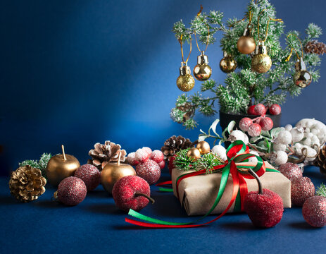 Christmas scene with handmade craft gift box with bow, decorative fir branch, balls and cones for tree decoraton. New Year winter holiday composition with presents, ornaments, decor on blue background