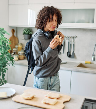 A teenage student eats a sandwich in a modern kitchen, wearing a backpack and headphones while preparing a quick breakfast before heading to school. The candid moment captures a casual morning routine
