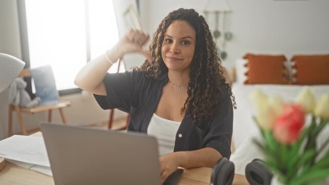 Woman at laptop giving thumbs down with hand visible in bedroom at a desk with papers headphones lamp and tulips; disappointment remote study.