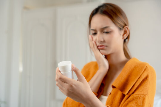 Toothache concept, woman holds her painful cheek while checking a medicine bottle at home, showing dental pain and self care, toothache, medication, pain, healthcare, treatment