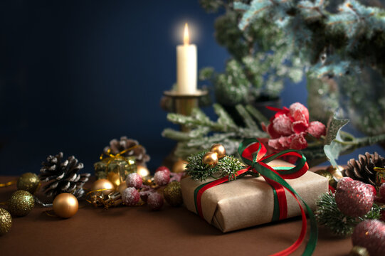 Christmas scene with handmade gift box with bow, candle, decorative fir branch, balls and cones for tree decoraton on blue background. New Year mysterious holiday composition with presents and decor.