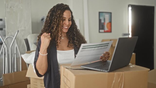 Woman reads a document beside a laptop on stacked moving boxes and raises fist in building while smiling; new home excitement.