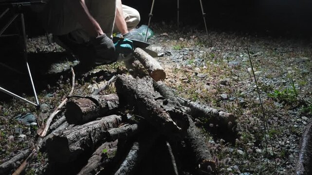 Person wearing protective gloves cutting a fallen birch tree log with a small cordless electric chainsaw, with sawdust flying around during nighttime yard work or land clearing activity