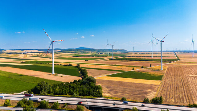Wind towers overlooking highway traffic and farm fields. Vehicles travel along a concrete transportation corridor bounded by cultivated vegetation.