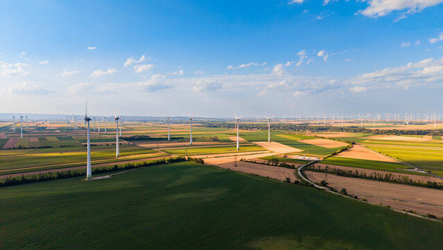Wind turbines standing in vast agricultural fields. Wide aerial shot of multiple wind turbines distributed across colorful cultivated farm fields under sky.