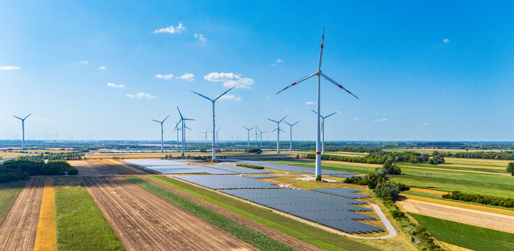 Wind turbines and solar panels in agricultural field. Aerial perspective of large solar panel array and wind turbines standing among green and brown fields.