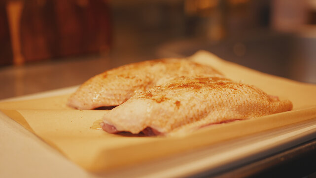 Close-up of raw seasoned duck breast skin-on fillets on parchment paper over a cutting board on a kitchen table. Preparation process of marinated meat before cooking in a warm home kitchen.