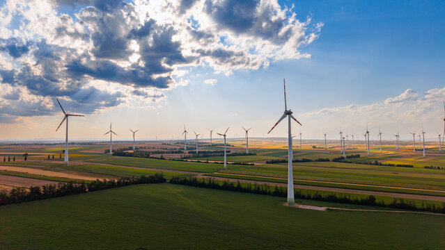 Wind power generators deployed across countryside farmland. High angle shot displays rotating tall pillars positioned between cultivated soil patches under scattered clouds.
