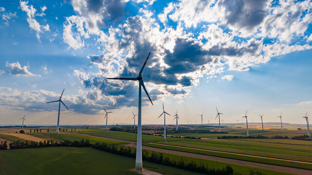 Wind turbine standing near agricultural land parcels. Drone camera records prominent tall energy tower with spinning blades overlooking flat terrain and scattered clouds.