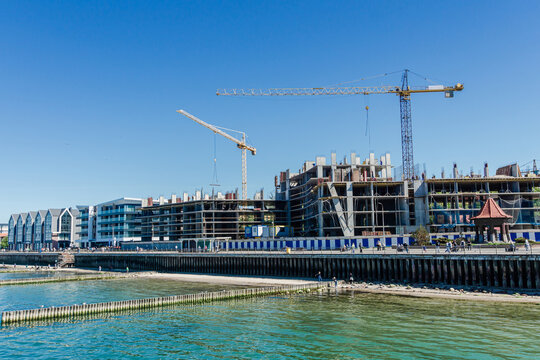 Active construction of residential apartment buildings along the seafront, with tower cranes rising above a coastal city skyline.