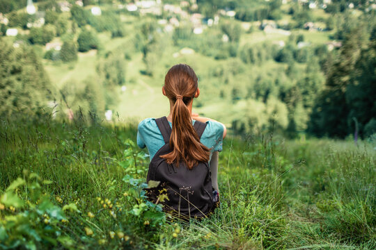 Woman hiker with backpack relaxing on mountain hillside overlooking green valley, travel, mindfulness, freedom, wellness and outdoor adventure concept
