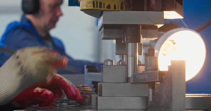Metal parts manufacturing on mechanical stamping press. Blue collar operator handles components beside stamping die and stacks of finished parts