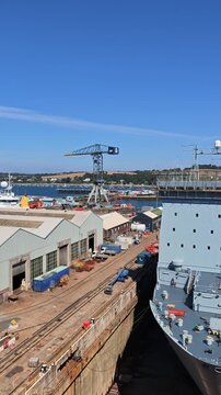 Falmouth Cornwall England UK. 21.08.2026. Video. A grey British warship sits in a dry dock for repairs with industrial cranes and shipyard buildings visible in the background on a sunny day