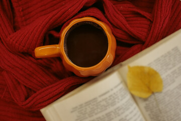 Fototapeta premium An orange mug of coffee resting on a red knitted sweater beside an open book and a yellow autumn leaf, with soft film grain, high quality photo